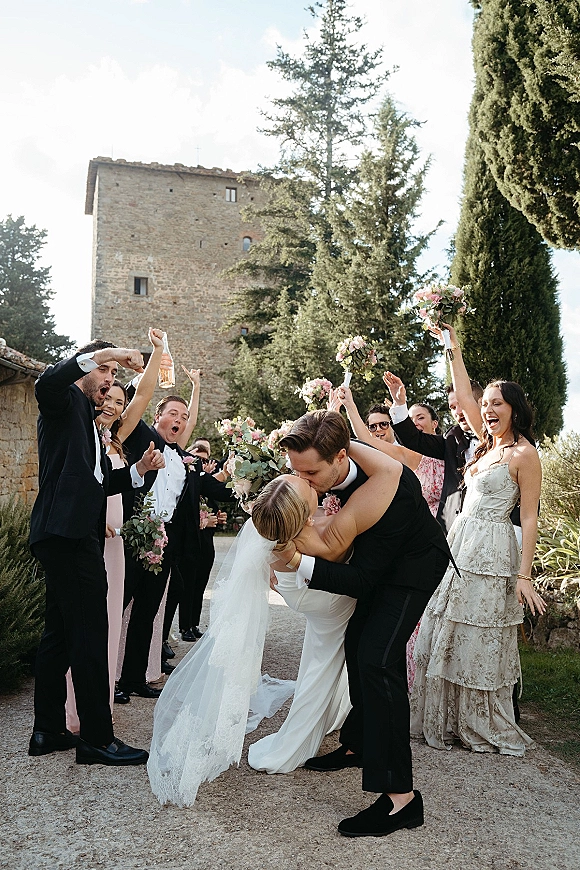 Wedding kiss as groom dips bride in veil and bouquet, wedding party cheering behind on a garden path by a stone building