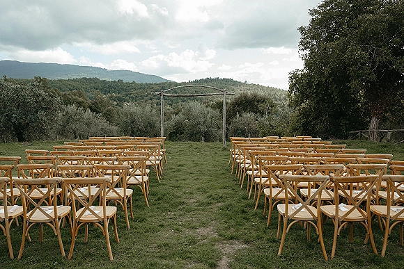 Ceremony setup for an outdoor wedding ceremony with wood crossback chairs lining the aisle to a rustic wooden arch on a lawn with mountains