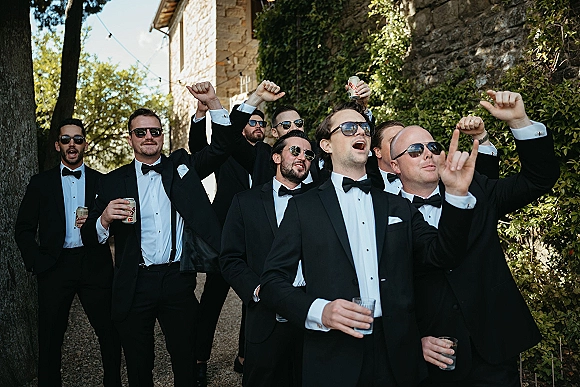 Groomsmen portrait with groomsmen group photo in black tuxedos and sunglasses, holding beer cans and rocks glasses by an ivy stone wall