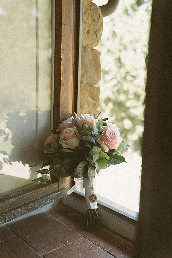 Bridal bouquet of pink roses with eucalyptus accent and white ribbon wrap, featuring a charm locket by a window in natural light
