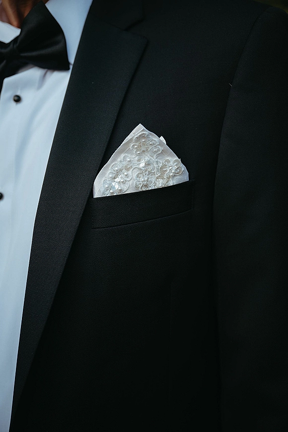 Groom tuxedo detail with satin lapel, black bow tie, and wedding pocket square in lace, plus shirt studs against a softly blurred backdrop