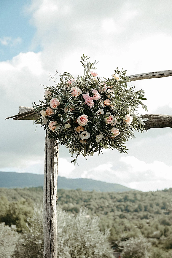 Wedding arch flowers arranged on a rustic wooden wedding arch with blush roses and greenery, set against mountains, trees, and blue sky