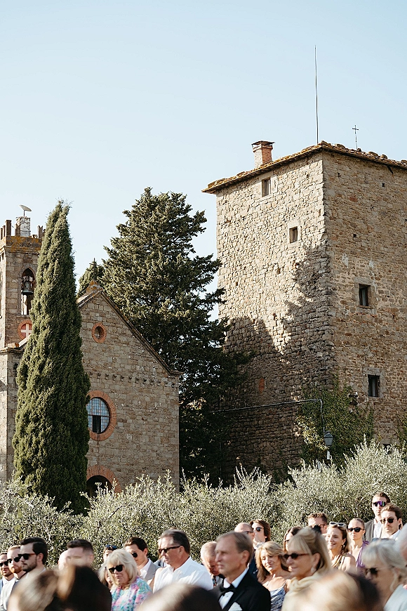 Outdoor wedding ceremony with wedding guests seated in sunglasses and suits, facing a stone church and bell tower under clear sky