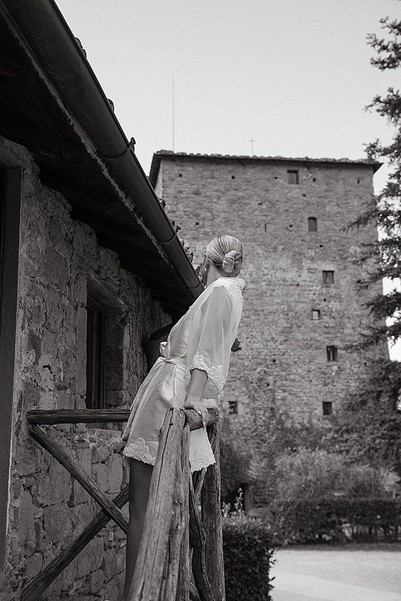 Bridal getting ready bride in lace robe with satin belt and earrings, hair in updo, standing by wooden railing near stone castle wall courtyard.