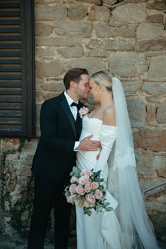 Wedding couple portrait of bride and groom embrace, touching foreheads as she holds a rose bouquet by an ivy-covered stone wall