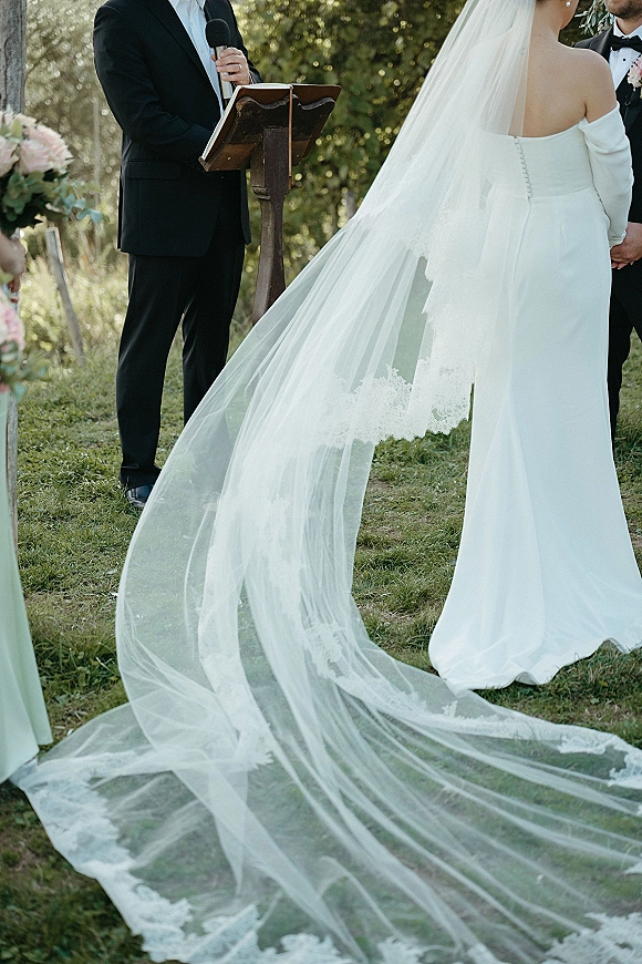 Ceremony moment as bride and groom hold hands at the altar, her long lace veil flowing, officiant speaking at a wooden lectern outdoors