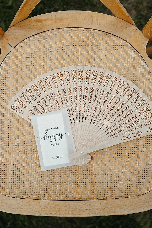 Happy tears kit with wedding happy tears packets beside a wooden folding fan on a woven cane chair against outdoor greenery
