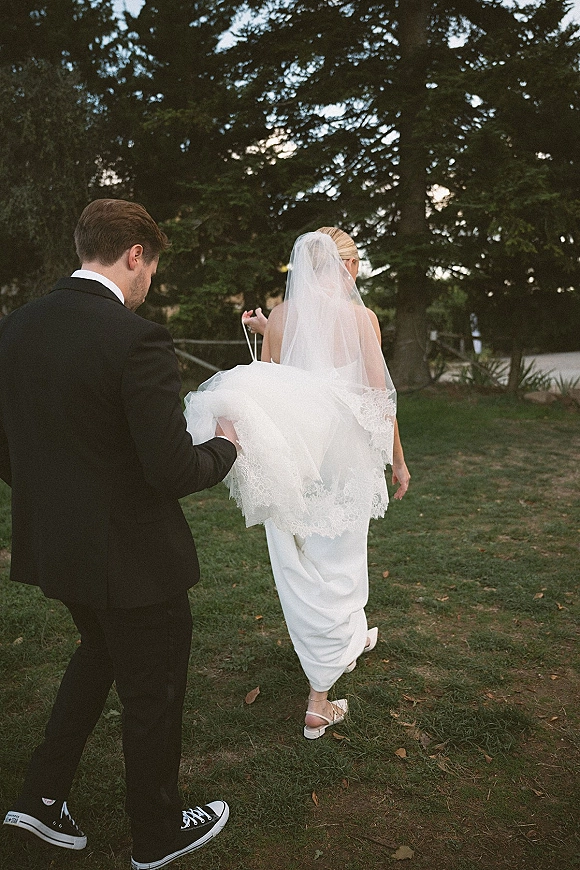 Wedding couple portrait of bride and groom walking away across a grassy lawn, long veil trailing, evergreens behind, groom in tuxedo and sneakers