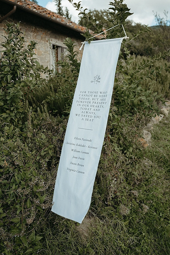 Wedding memorial sign, in memory wedding sign on a hanging fabric banner with rope and greenery, set by garden shrubs near a stone building