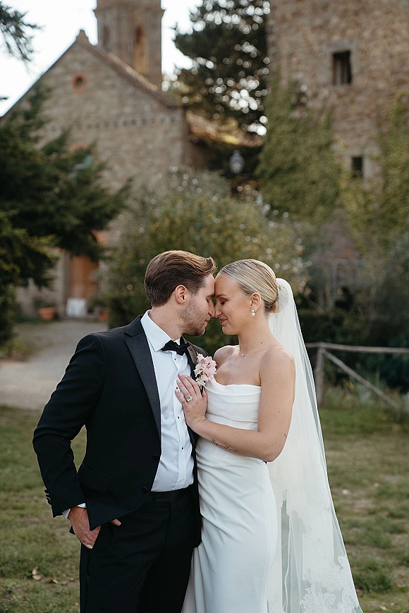Couple portrait of bride and groom embrace with a forehead kiss, her veil flowing beside a stone church and garden pathway backdrop
