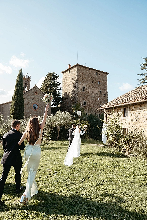 Wedding recessional as bride walking away with bouquet and long veil beside groom, bridesmaid cheering with bouquet outside a stone church courtyard
