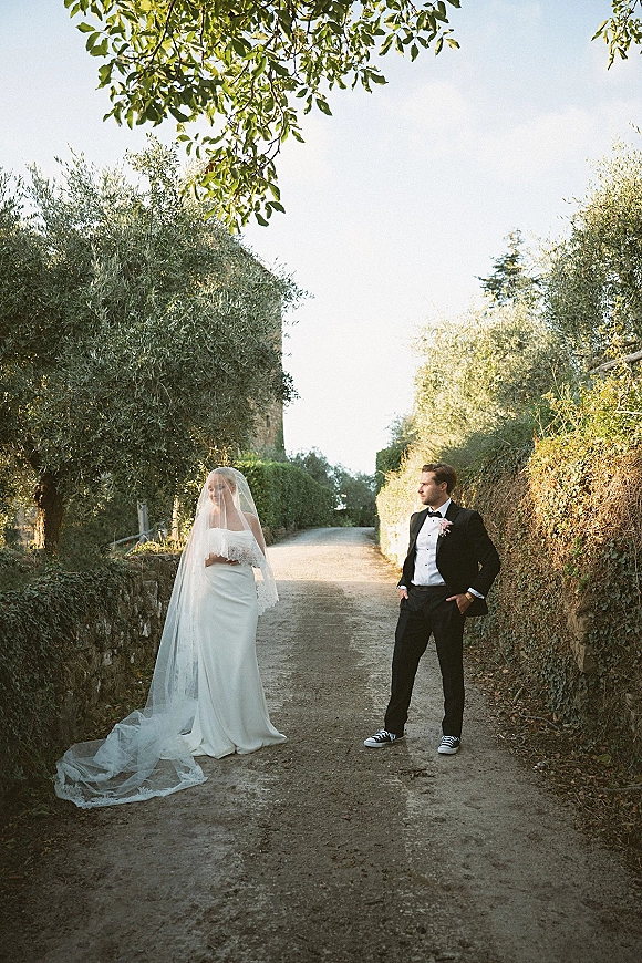 Couple portrait of bride and groom portrait with long lace-trimmed veil and black tuxedo, standing on a tree-lined dirt road by a stone wall