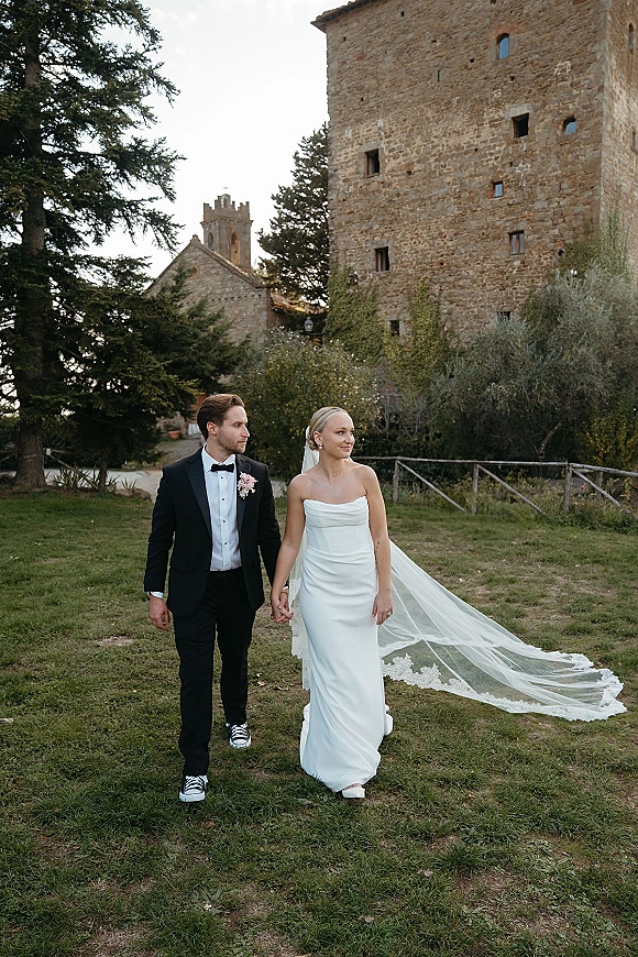 Couple portrait of bride and groom walking hand in hand, her long lace veil flowing beside a stone building and rustic fence