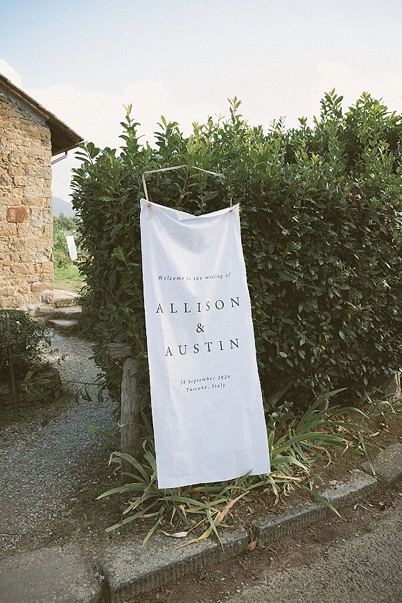 Wedding welcome sign on a fabric banner with rope on a hanging frame, set by a stone building and green hedge along a gravel path