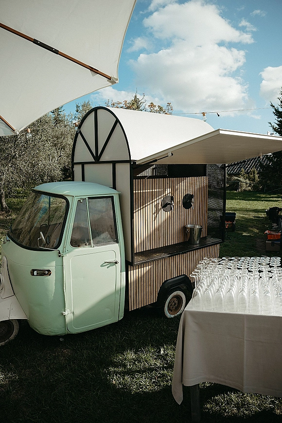 Wedding bar setup with a mobile wedding bar truck, wine glasses and linens under a parasol on a grassy lawn with string lights and trees