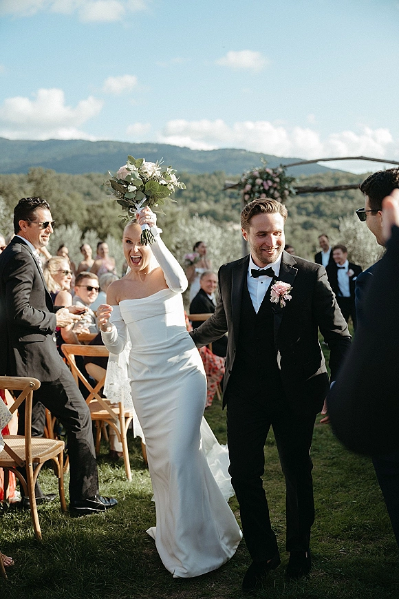 Wedding recessional as bride lifts her bouquet beside groom in tux, walking past cheering guests on a lawn aisle with mountains beyond