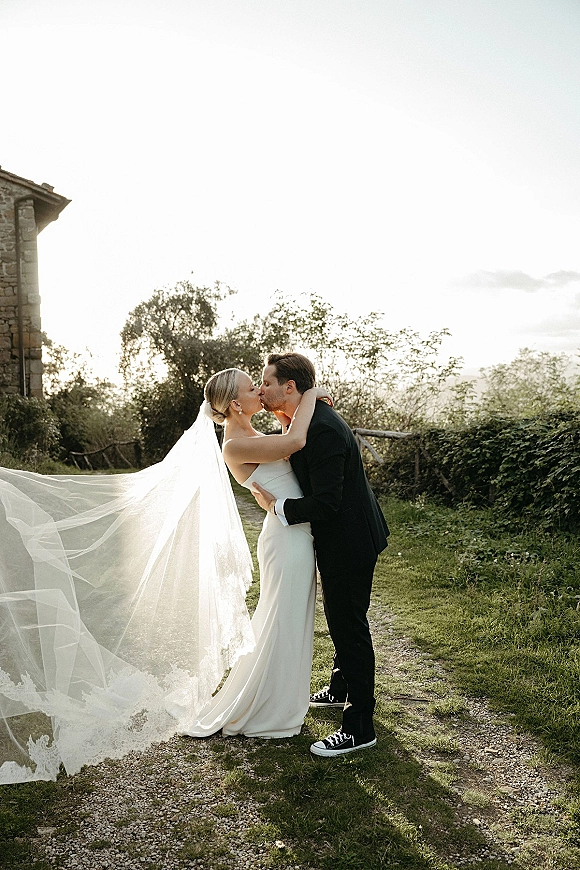 Wedding kiss portrait of bride and groom kissing, her long veil blowing as they embrace on a grassy path by trees and stone building