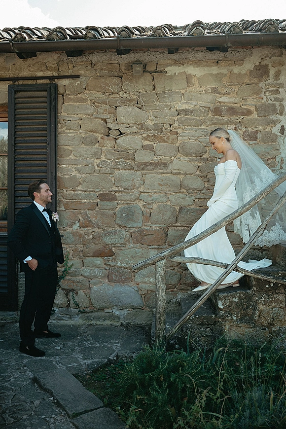 First look moment as bride walking down stairs in an off the shoulder wedding dress with veil toward groom in tux by a rustic stone wall