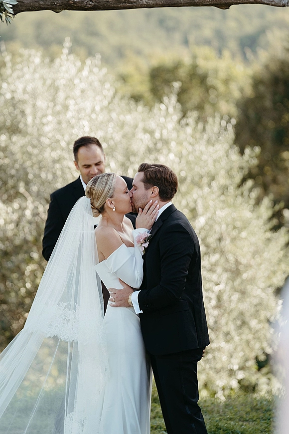 Wedding kiss at an outdoor ceremony as bride in off-the-shoulder dress and veil holds groom’s face in tuxedo before greenery hillside