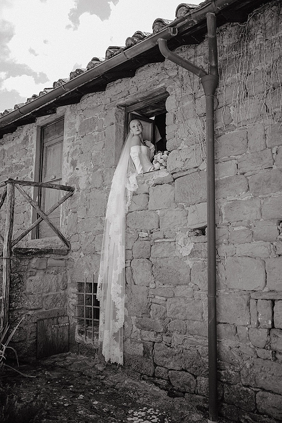 Bridal portrait of a bride with long veil holding a bouquet, leaning out a window of a rustic stone building with ivy vines