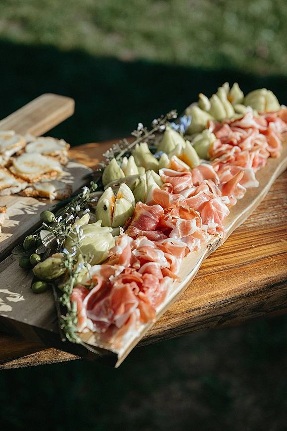 Charcuterie board for wedding cocktail hour food with prosciutto, artichoke hearts, olives, herbs and crackers on a wooden board outdoors