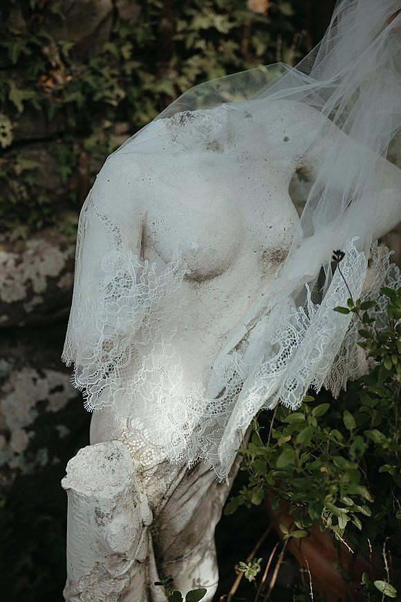 Bridal veil close-up showing lace edge veil trim on a mannequin bust, sheer tulle draped against greenery, stone wall, and terracotta planter