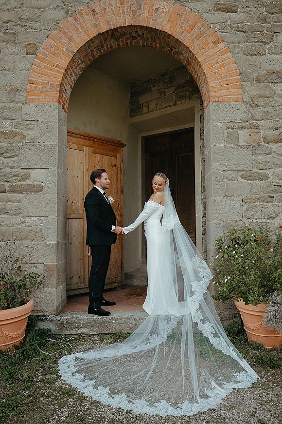 Couple portrait of bride and groom holding hands, her long lace cathedral veil flowing by a stone archway and rustic wooden door