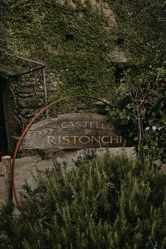 Wedding venue sign on a rustic wooden panel inside a metal hoop, decorated with ivy against a vine-covered stone wall backdrop