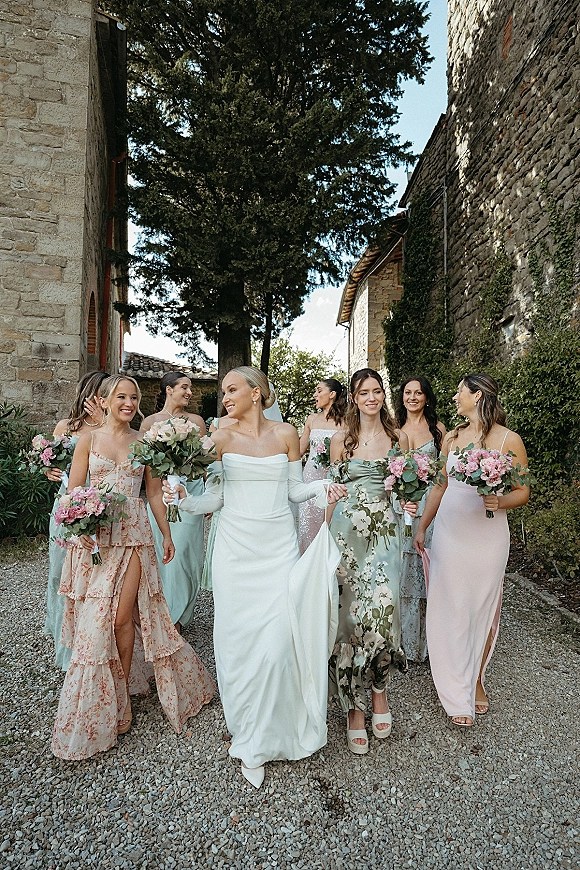 Bride with bridesmaids walking together in mismatched dresses, holding pink bouquets, her veil flowing beside ivy-covered stone buildings
