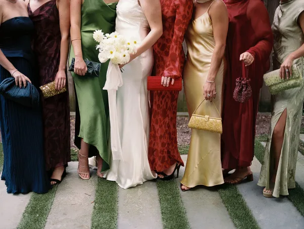 Bridesmaid dresses in mismatched satin jewel tones with clutches and heels, posed along a stone walkway beside a garden wall