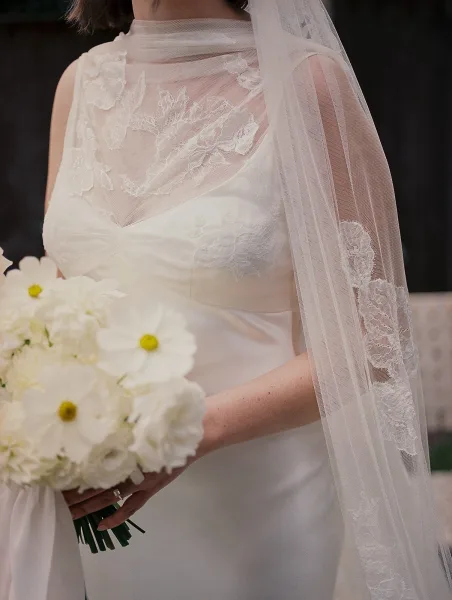 Bridal dress detail with illusion neckline and lace applique, bride holding a white bouquet and wedding ring on an outdoor walkway