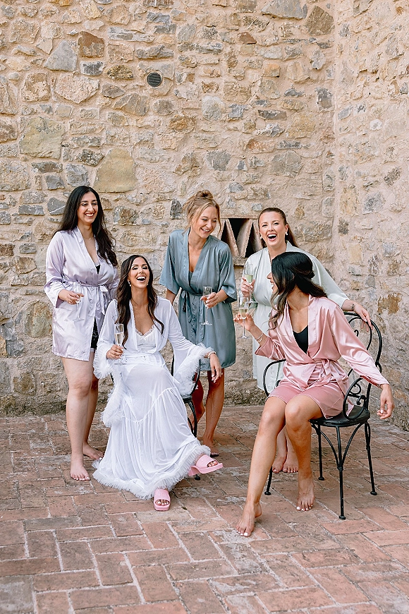Bridesmaids getting ready in bridal party robes, raising champagne flutes and laughing beside a stone wall on a brick patio