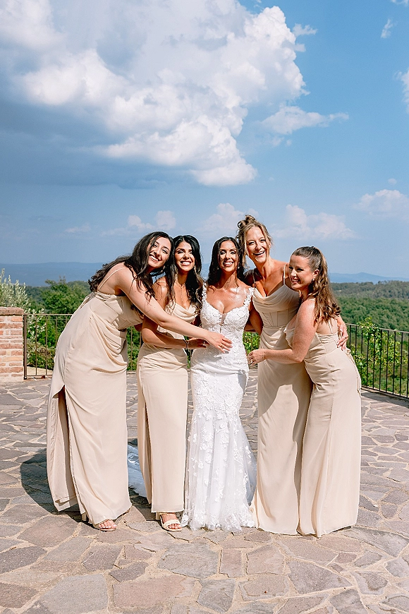 Bridesmaids photo of the bride with bridesmaids hugging her in champagne dresses on an outdoor terrace with mountain views under blue sky