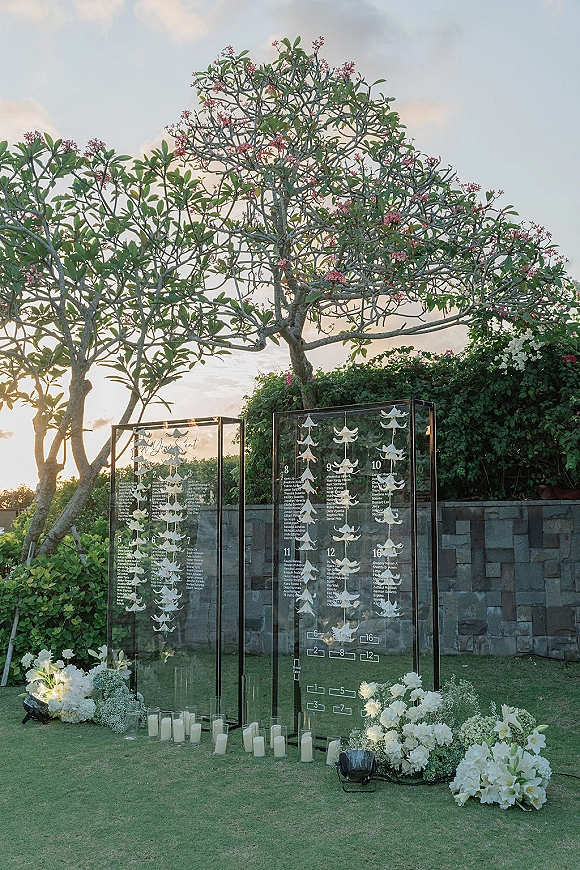 Wedding seating chart on clear acrylic panels with white butterfly escort cards and candles, set on a garden lawn by stone wall and trees