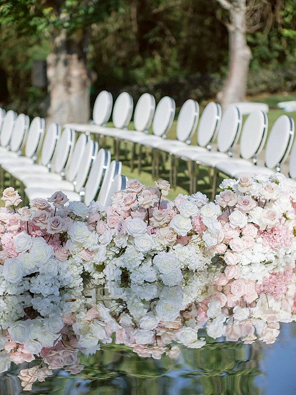 Ceremony aisle decor with curved rows of white chairs and low rose and hydrangea arrangements beside reflective water on a garden lawn