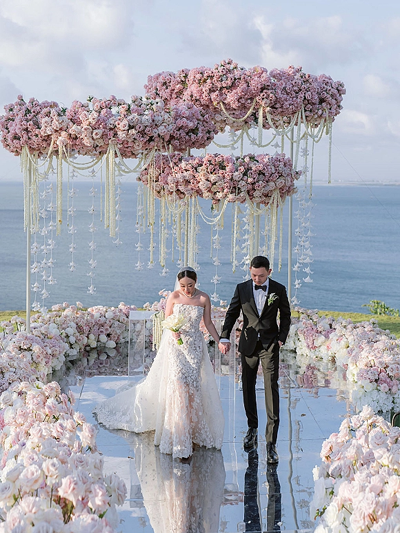 Wedding recessional with bride and groom walking aisle beneath a pink floral arch, mirrored walkway and ocean view beyond horizon