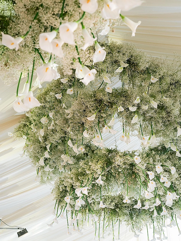 Hanging floral installation with wedding ceiling flowers, calla lilies, baby's breath, and greenery beneath a white draped tent canopy