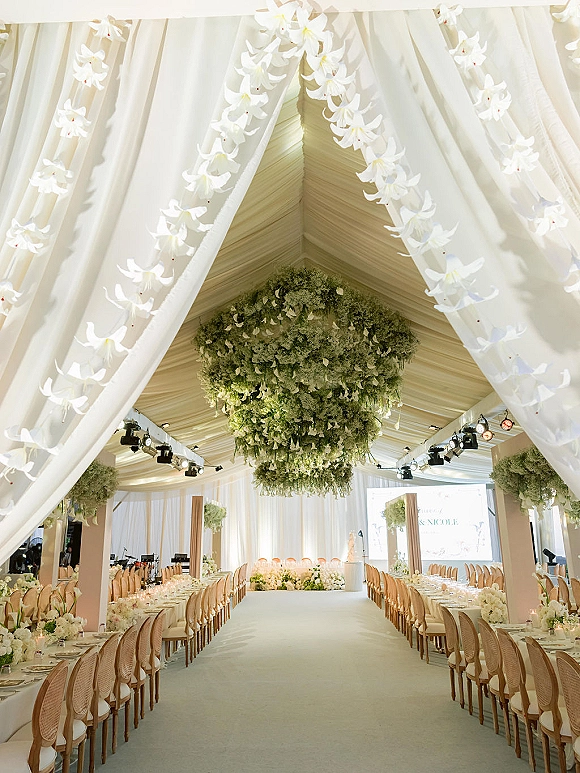 Reception tablescape with long banquet tables, rattan chairs, candles and white florals beneath a white draped ceiling and hanging greenery installation
