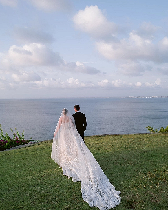 Couple portrait of bride and groom from behind, her lace train and veil flowing as they face the ocean horizon on a cliff lawn