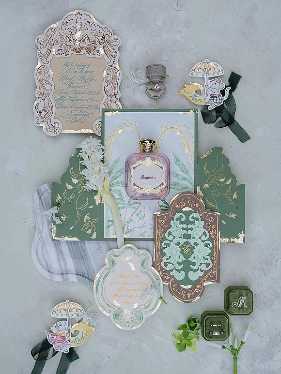 Invitation suite wedding invitation flat lay with green and gold cards, rings, ribbon, perfume, and flowers on a gray stone surface