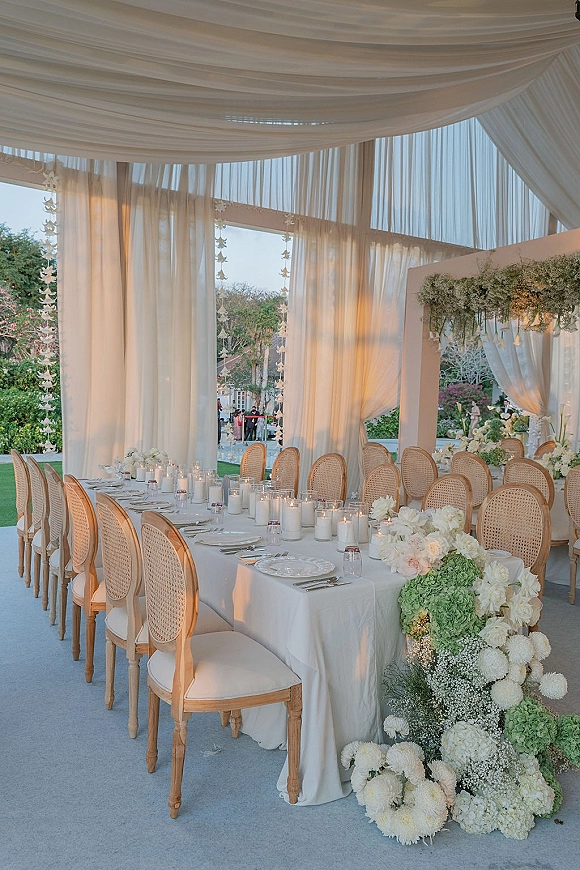 Reception tablescape with long banquet table decor, white linens, taper and hurricane candles, white and green hydrangea garland under draped canopy in garden.