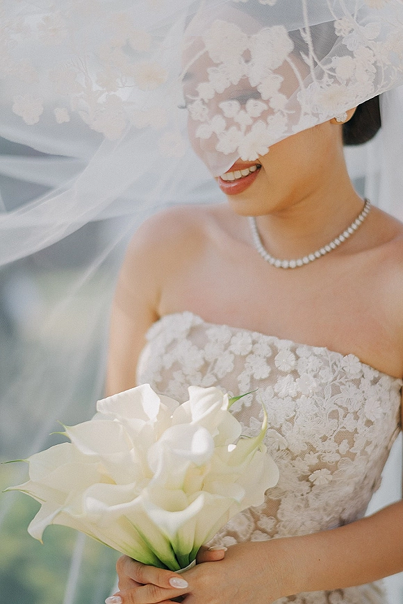 Bridal portrait of a bride holding bouquet of white calla lilies, smiling under a lace veil with pearl necklace on a soft neutral backdrop