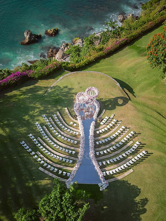 Ceremony setup with aerial wedding ceremony seating in a curved semicircle, mirror aisle runner to a floral arch on an oceanfront lawn