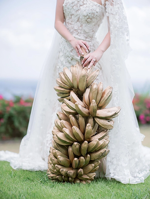 Bridal portrait of a bride holding bananas like a bouquet, showing lace wedding dress, veil, and engagement ring on a grassy lawn