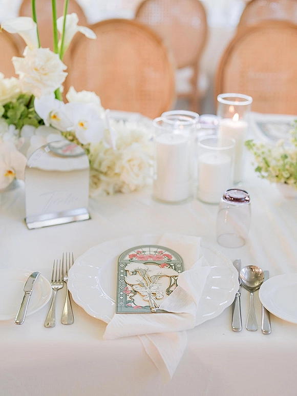 Reception tablescape with wedding place setting, white florals and taper candles in glass votives on a white tablecloth in a reception hall