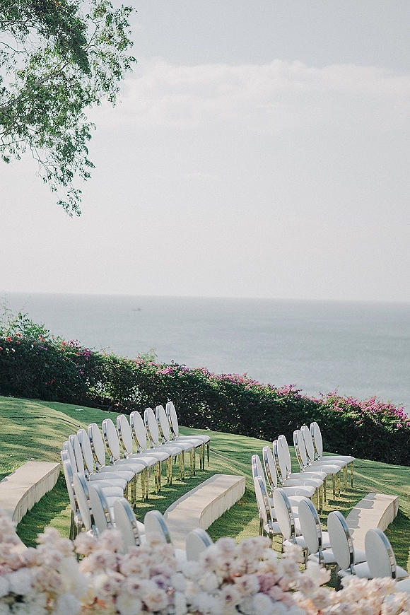 Ceremony seating with outdoor ceremony seating in a U-shaped layout of white chairs and aisle runner facing a floral hedge by the ocean view