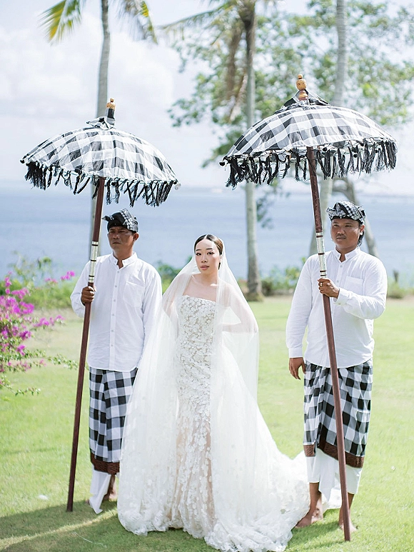 Bridal portrait of a bride with veil in a strapless lace wedding dress, with attendants holding parasols by ocean and palm trees