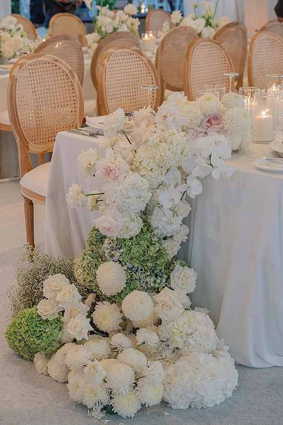 Reception tablescape with wedding head table flowers spilling across a white tablecloth, with glass cylinder candles, place settings, and rattan chairs indoors