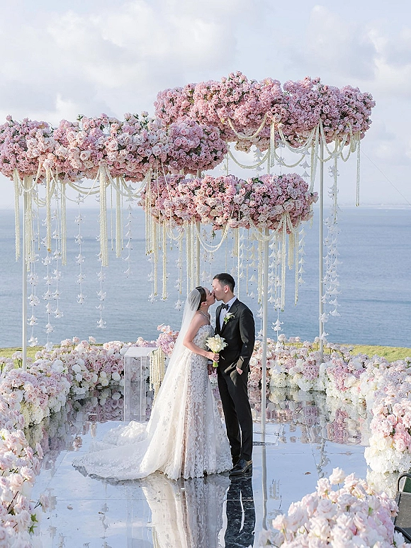 Wedding kiss with bride and groom kissing under a pink rose floral arch, veil and tux reflected on a mirrored aisle by the ocean