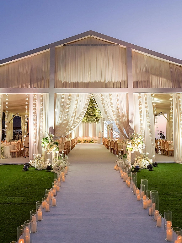 Ceremony aisle decor with a candlelit wedding aisle, white draping and hanging floral garlands under a tent on the lawn at dusk
